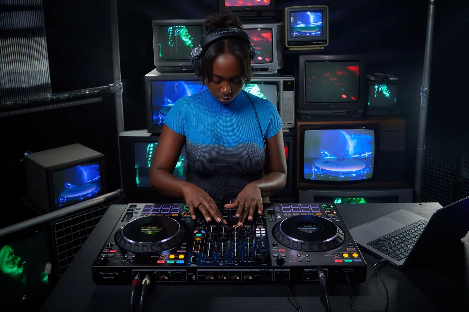 A DJ playing a Pioneer DJ DDJ-FLX10 in a room stacked with old televisions